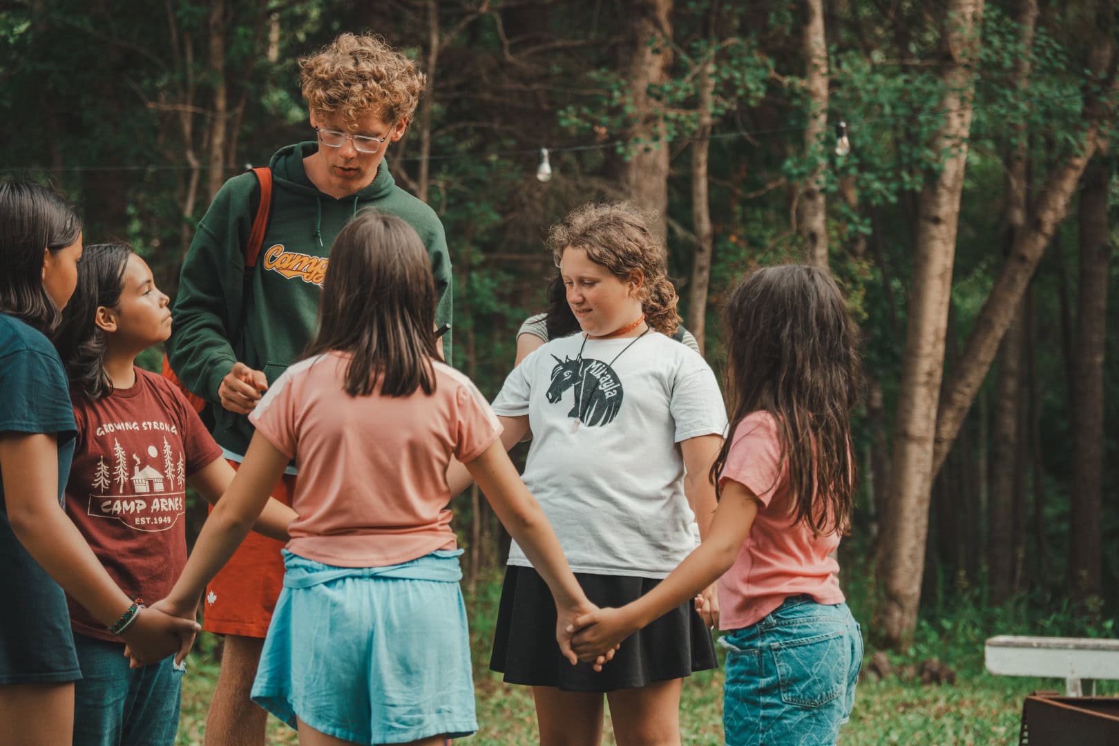 Children dressed casually, holding hands in a circle during outdoor camp activities at Camp Arnes in a forest setting.