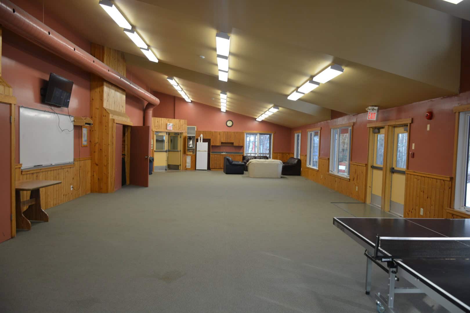 Spacious indoor recreation room at Camp Arnes featuring comfortable seating, large windows, ping pong table, whiteboard, and accessible amenities in a rustic setting.