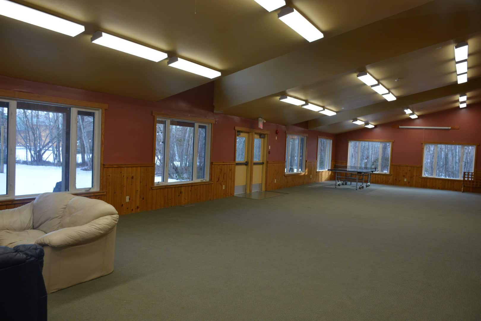 Indoor recreation hall with large windows showcasing outdoor snowy landscape, wood paneling, and ceiling lighting at Camp Arnes.