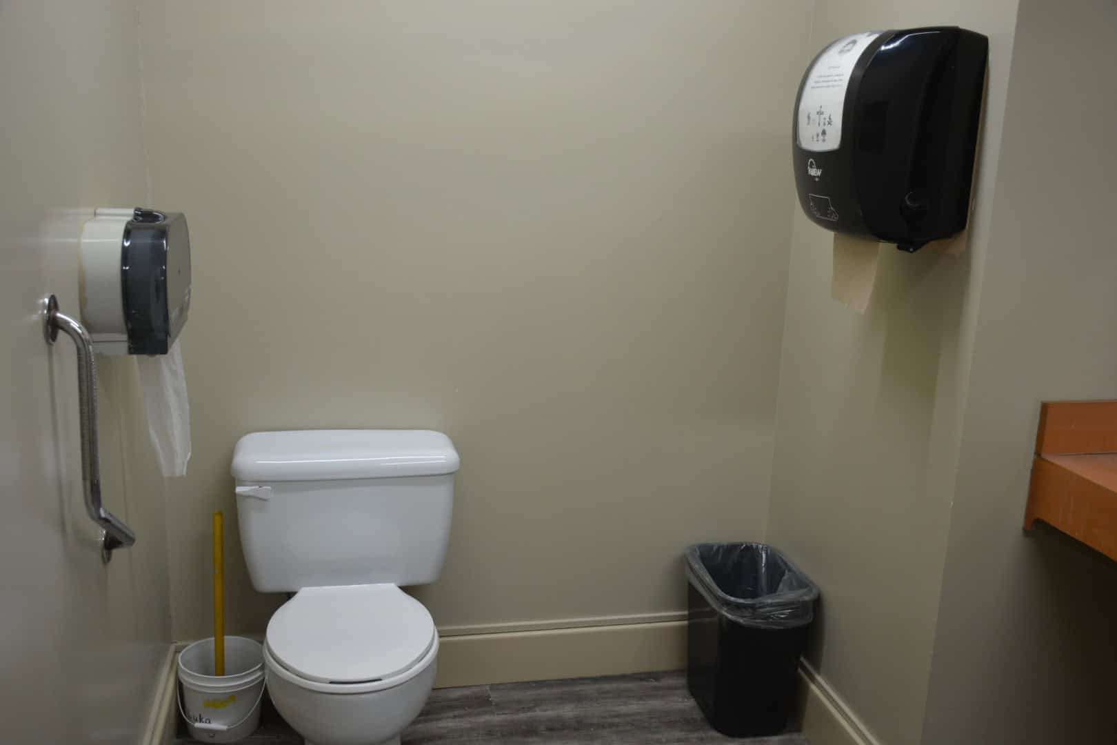 Toilet with handrail and paper towel dispenser in a camp facility bathroom.