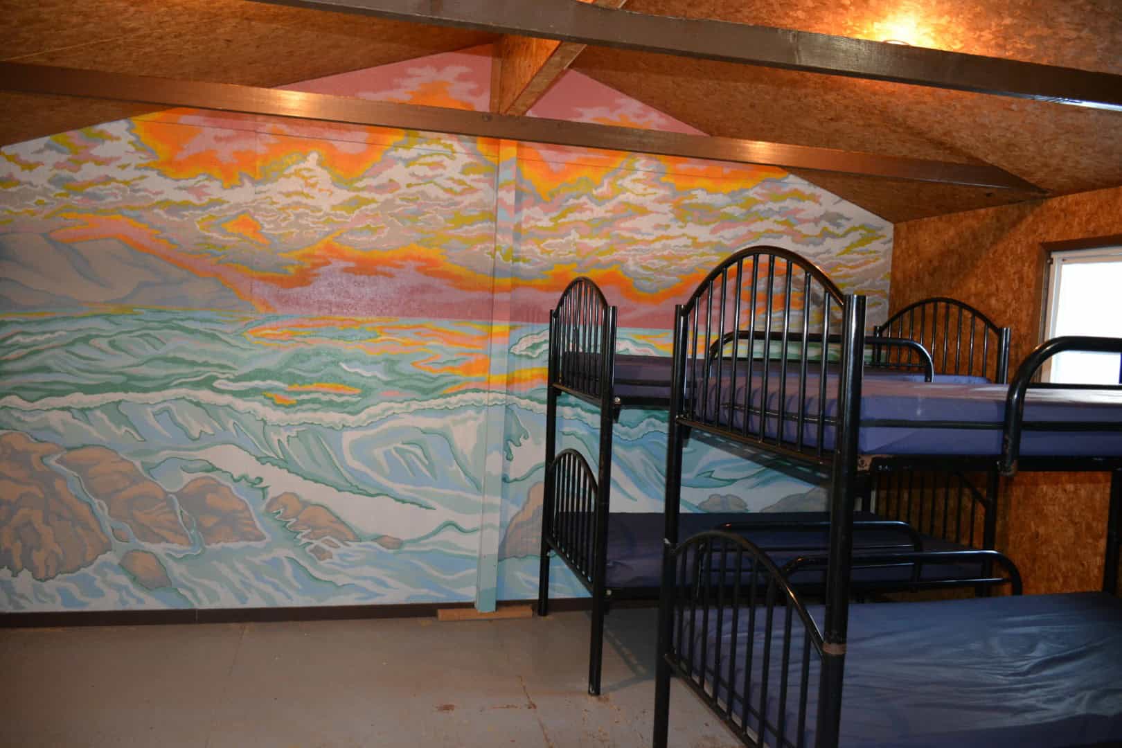 Colorful ocean wave mural inside a rustic cabin at Camp Arnes, featuring bunk beds with black metal frames under a wooden ceiling.