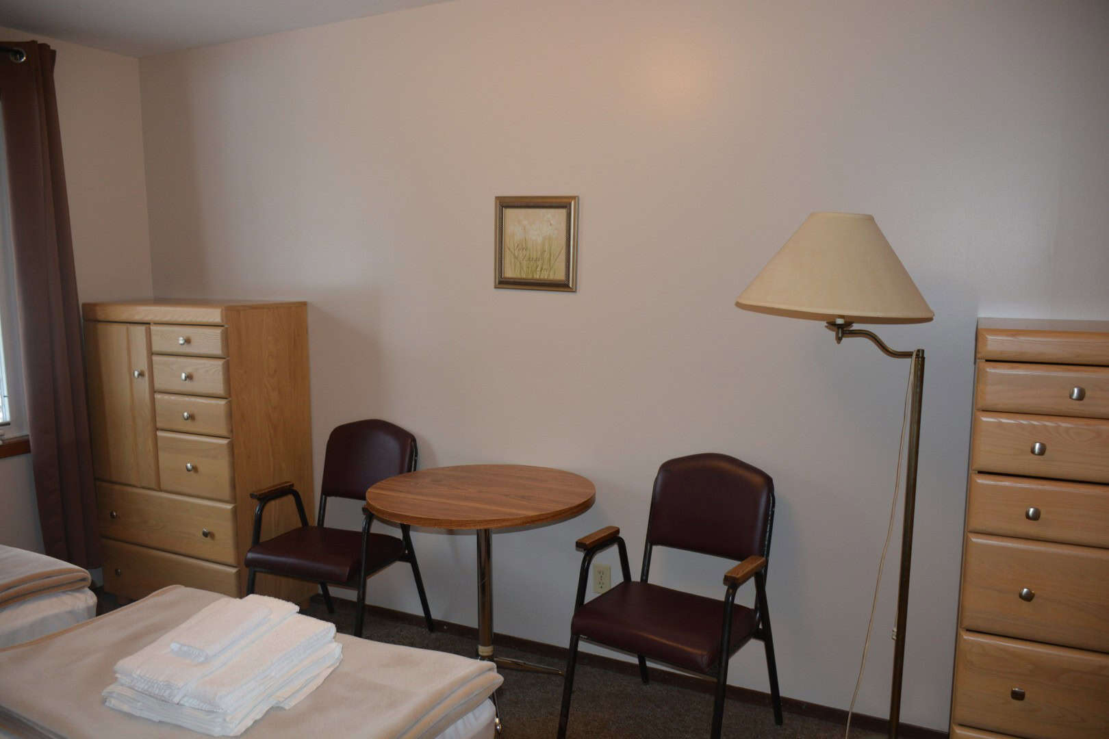 Cozy dormitory-style room at Camp Arnes with wooden furniture, chairs, a small round table, and neatly folded towels on a bed, offering comfortable accommodations near outdoor activities.