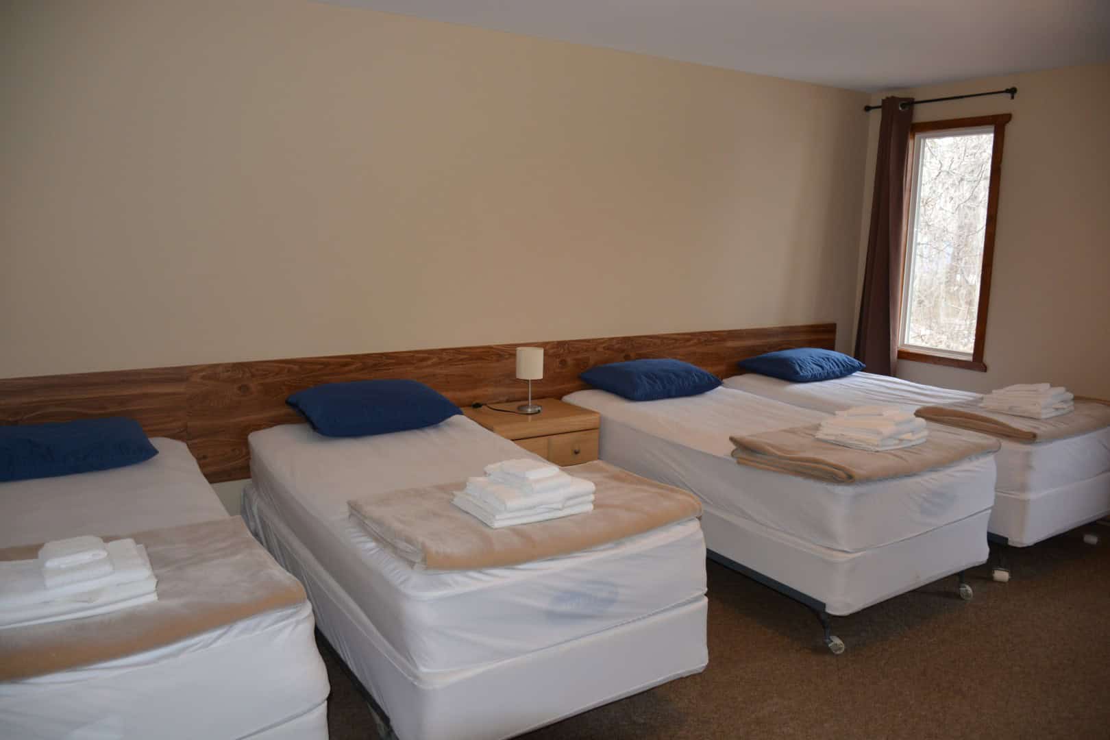 Cozy hotel room with four neatly arranged beds, blue pillows, and a wooden headboard at Camp Arnes. Natural light streams through a large window, creating a welcoming atmosphere for guests.
