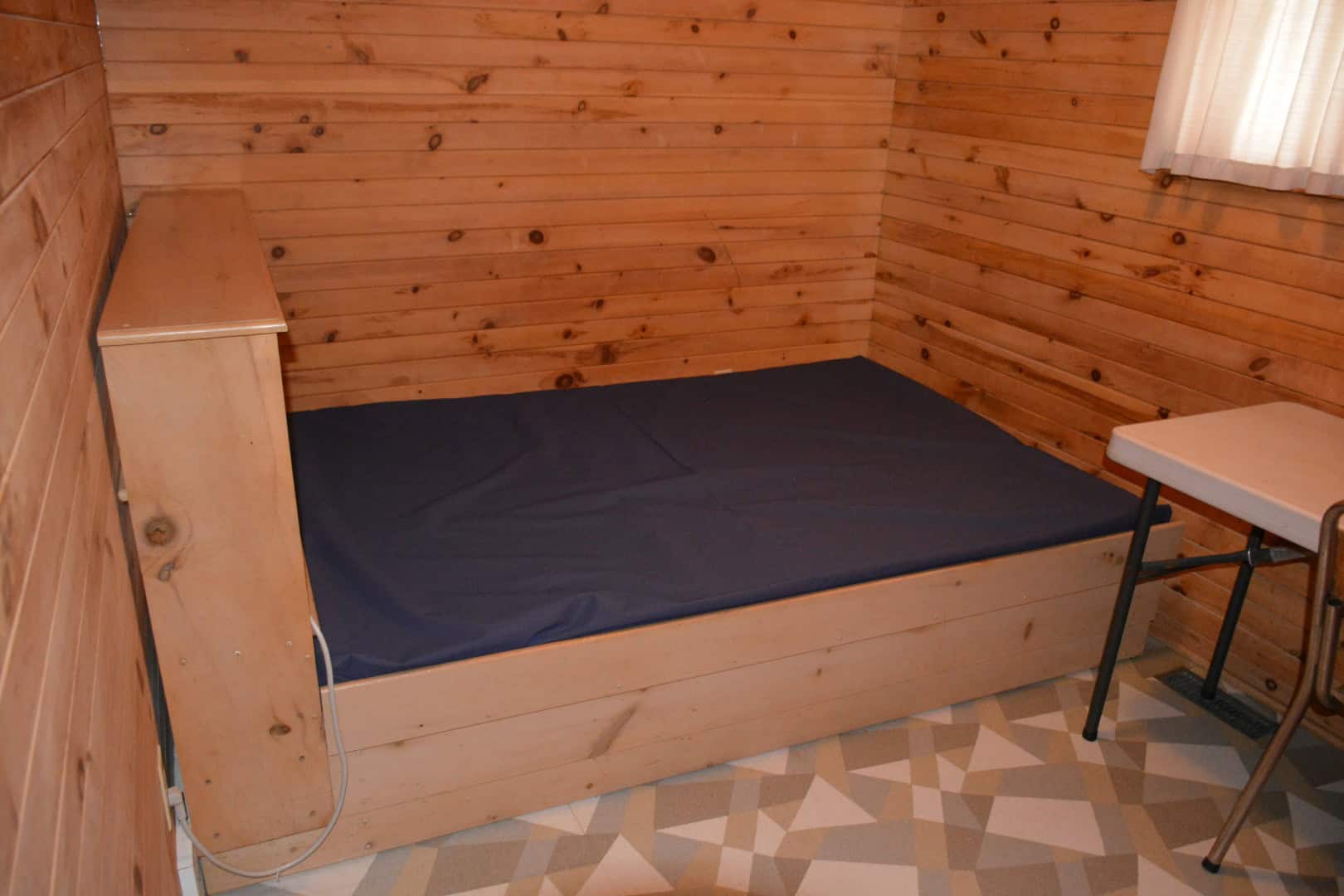 Cozy wooden cabin bedroom at Camp Arnes, featuring a wooden bed frame with a navy blue mattress, a small side table, and warm wood-paneled walls in a tranquil outdoor retreat setting.