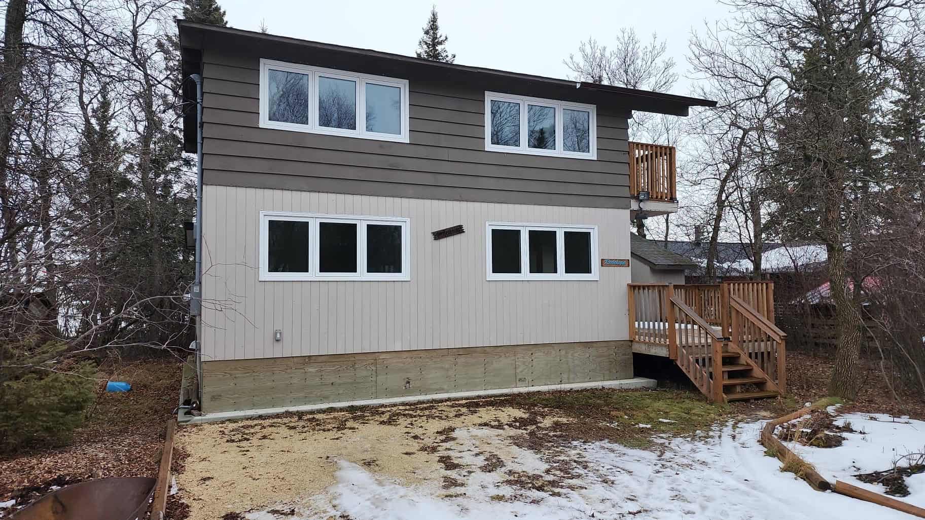 Modern two-story cabin at Camp Arnes with gray and beige siding, surrounded by winter trees, featuring a wooden staircase and small balcony, perfect for outdoor camping and retreat experiences.