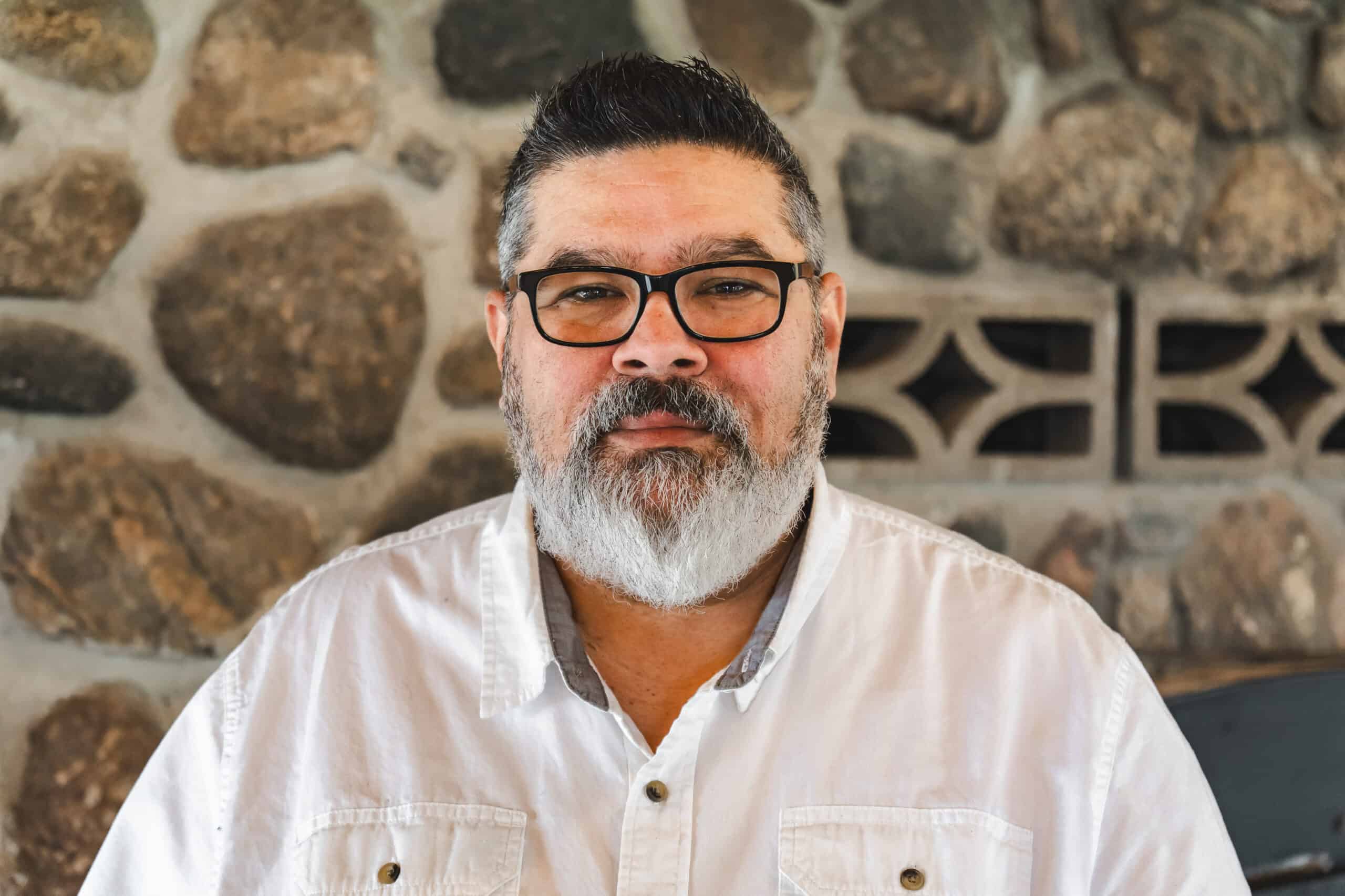 Relaxed man with glasses and a grey beard posing indoors against a rustic stone wall, representing family-friendly outdoor accommodations and camping experiences at Camp Arnes.