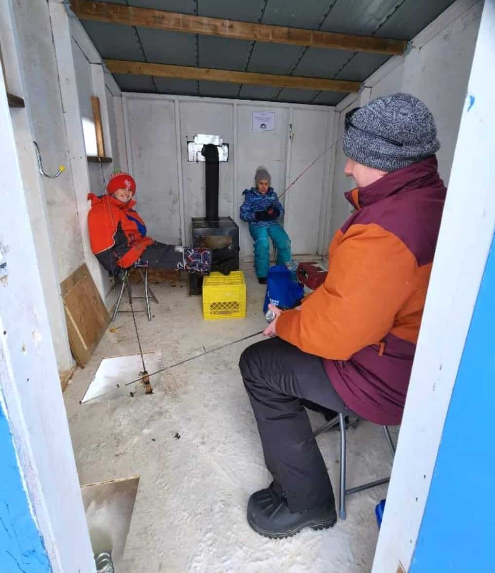 Fishing inside a cozy cabin at Camp Arnes, winter outdoor adventure, family enjoying ice fishing, warm clothing, snow-covered landscape, rustic cabin setting, outdoor recreation at Camp Arnes.