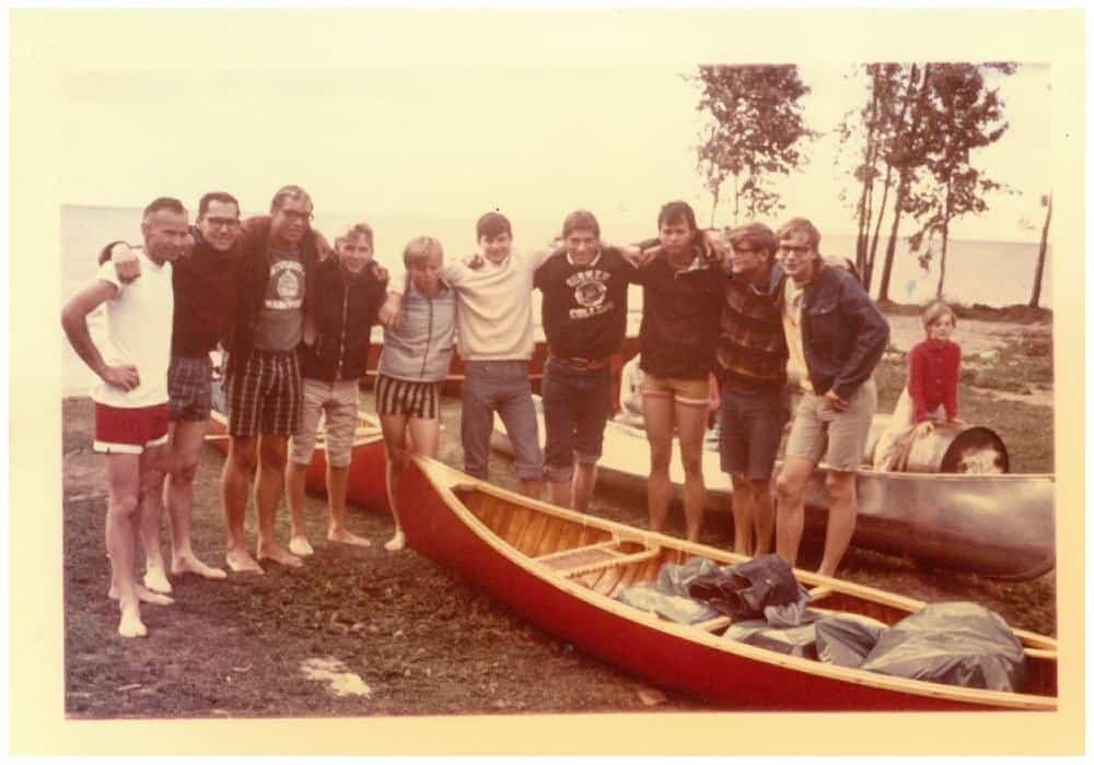 Older photo of a group of kids and teenagers at Camp Arnes, standing beside canoes on a grassy shore, showcasing outdoor camp activities, friendship, and nature.